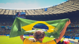 homem de costas segurando uma bandeira do brasil erguida com as duas mãos em estádio de futebol, com céu azul