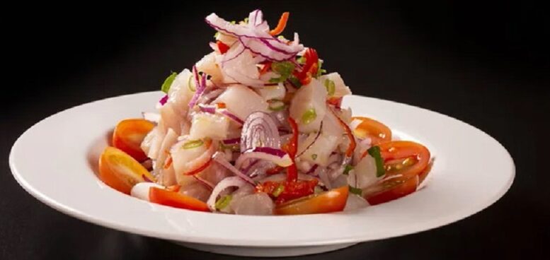 imagem de um prato de ceviche em fundo preto. é um rpato fundo de louça branca, com um ceviche montado como 'morro', cercado de tomatinhos cereja partidos ao meio