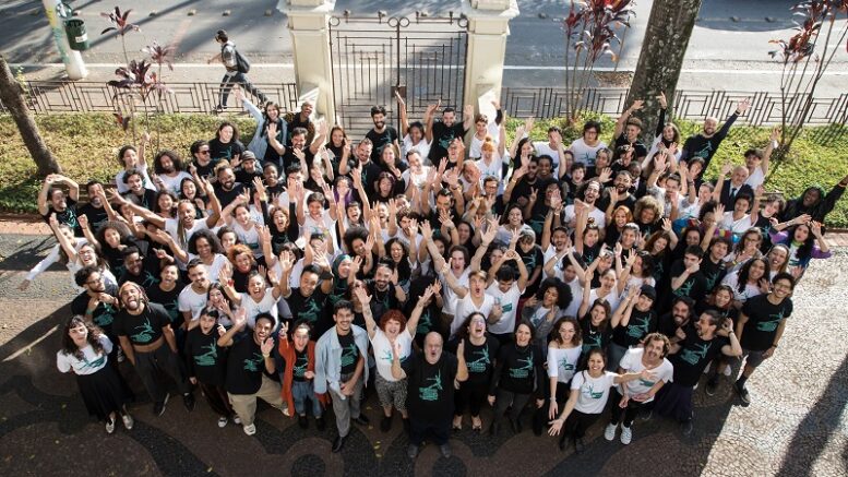 Alunos e equipe da São Paulo Escola de Dança vistos de cima, possivelmente de uma janela da escola. Eles estão no Jardim, olhando para a foto com os braços erguidos e sorrisos largos.