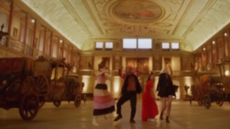 em um salão de museu com carruagens e afrescos no teto, 4 personagens dançam lado a lado vistos de longe.