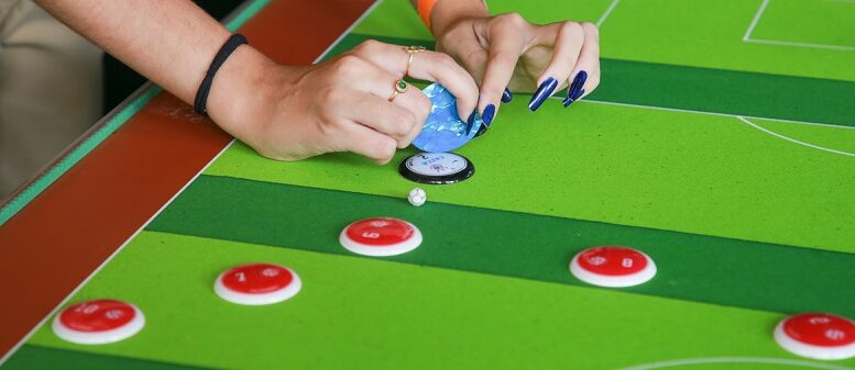 mãos de uma mjlher com unhas enormes azuis jogando futebol de botão, campo listrado de verde e o time dela parece ser um botão preto, contra botções vermelhos que aparecem em maioria na imagem.