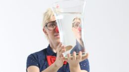 homem de cabelos platinados com óculos de grau e nariz de palhaço segura um vaso de vidro cheio de água em frente ao rosto,