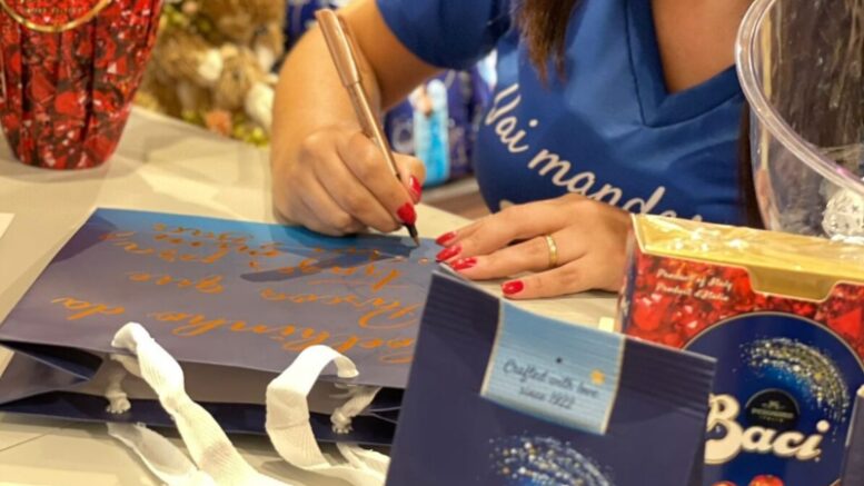 detalhes das mãos de uma calígrafa personalizando sacolas de papel azul, em primeiro plano uma sacola de ovo de pascoa da marca baci perugina