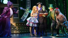 cena no palco com as duas bruxas checando um livro ao lado do Mágico de Oz, um ser meio gente meio bicho e outra personagem e observadas do outo lado do palco por outra peersonagem feminina.