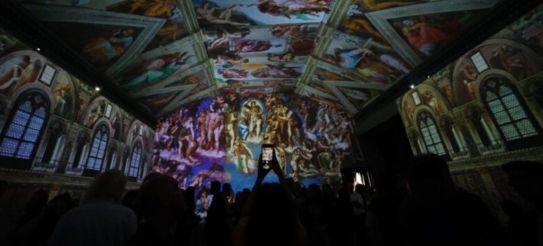 imagem de sala da exposição de Michelangelo, escura com o teto ilminado e é possível ver que o chão está lotado com muitos celulares fazendo fotos