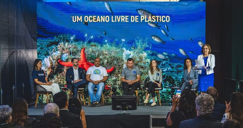 imagem do lançamento da campanha Pare o Tsunami de Plástico, com pessoas em um palco baixo, uma delas em pé falando ao microfone, as demais sentadas em cadeiras. no fundo uma foto do oceano com a frase 'uma oceano livre de plástico'.