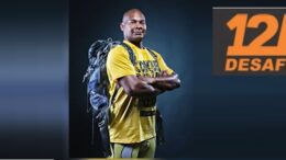 Carlos Dias de camiseta amarela, braços cruzads e uma mochila nas costas, há uma logo do desafio das 12h.