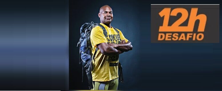 Carlos Dias de camiseta amarela, braços cruzads e uma mochila nas costas, há uma logo do desafio das 12h.