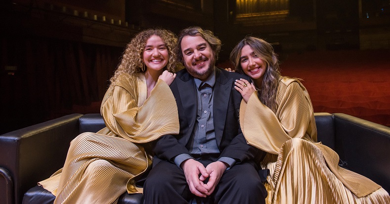 Ana Caetano e Vitória Falcão com vestidos em tons de dourado fosco, sentadas no chão do palco com o maestro Rodrigo Toffolo