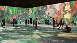 Imagem de um grande salão com projeção de imagens de flores de Van Gogh nas paredes e chão e diversas pessoas andando ou sentadas no chão admirando.