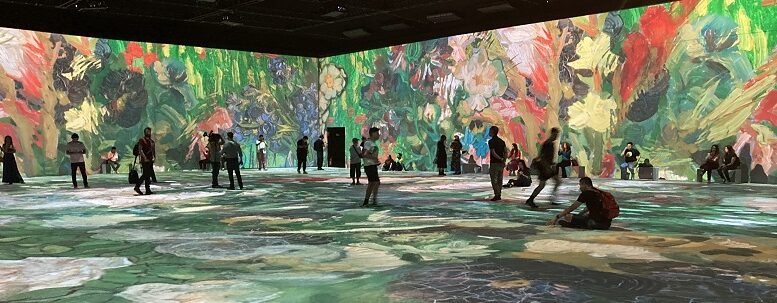 Imagem de um grande salão com projeção de imagens de flores de Van Gogh nas paredes e chão e diversas pessoas andando ou sentadas no chão admirando.