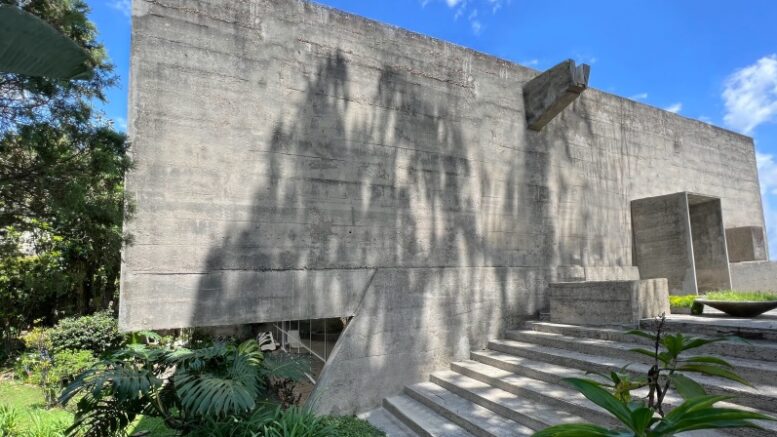 Frente da Casa Miani, projeto em concreto sem janelas voltadas para frente e uma escada larga com degraus baixos,