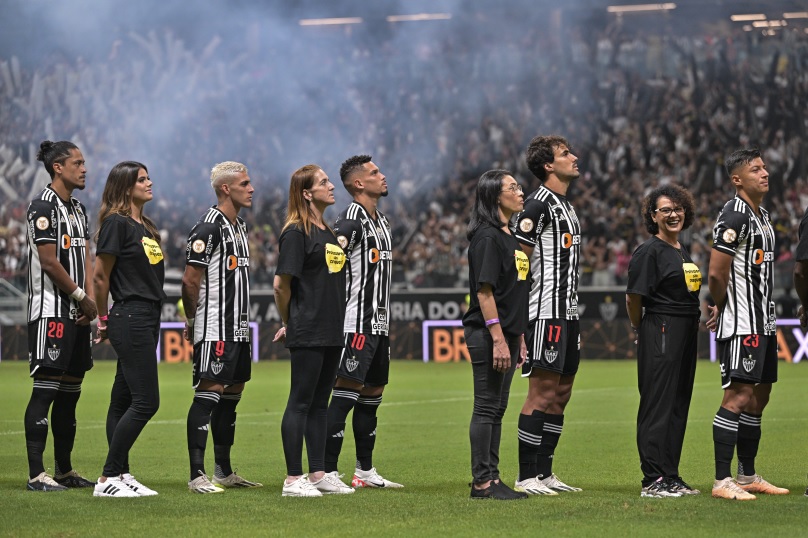 imagem de jogadores de futebol do Atlético Minieiro perfilados ao lado de professoras no gramado do Mineirão. 