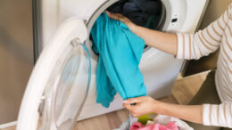 imagem de roupas sendo colocadas dentro de uma máquina de lavar, os braços paracem ser de uma mulher. A abertura da máquina de lavar é frontal.