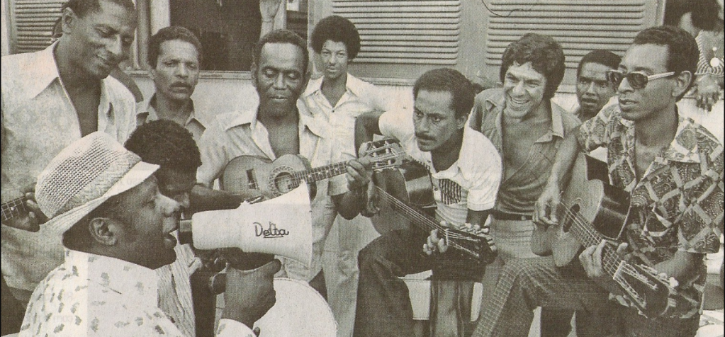 imagem em preto e branco de uma roda de samba e o mestre Candeia cantando com um megafone.
