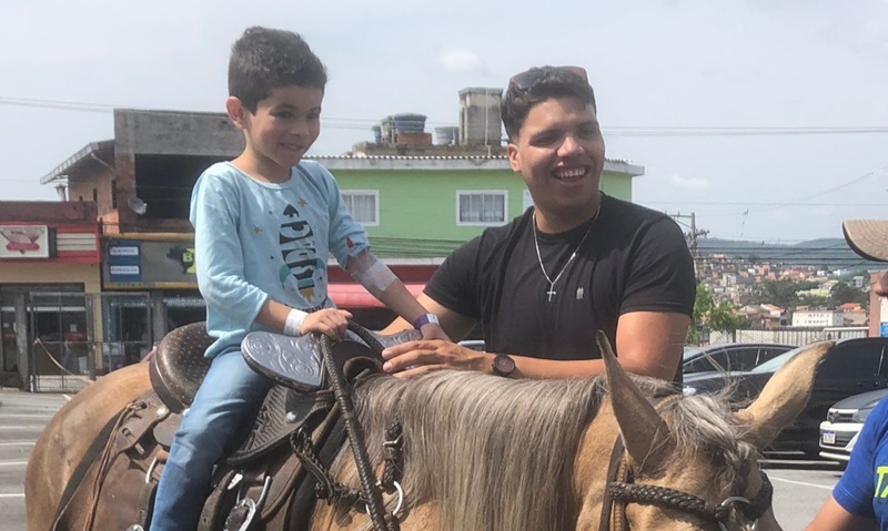 menino sorrindo em cima da égua, ele está sem máscara mas ainda tem o acesso sanguíneo no braço. há um instrutor sorrindo, em pé ao lado do cavalo, apoiando o garoto.