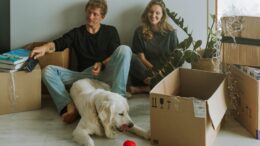 um casal sentado no chão de uma sala, com várias caixas de papelão de mudança e um cachorro golden com deitado em frente a eles com uma bolinha vermelha
