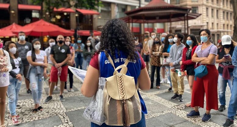 imagem de um grupo de turistas em roda, todos usando mascaras, olhando para uma guia que está de costas para a foto, com uma mochila e uma prancheta e a identificação bordada no colete.