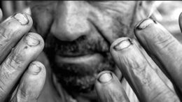imagem em preto e branco das mãos de um trabalhador, quase sem unhas, elas estão sendo mostradas junto ao rosto, do qual se vê do nariz para baixo, pele marcada, barba.