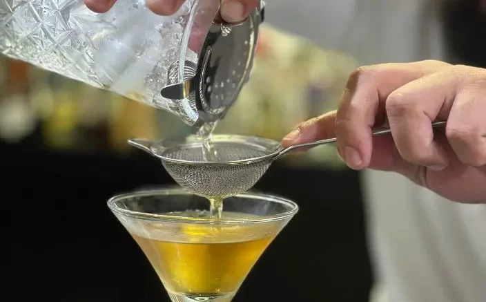 mãos de um bartender coando algum liquido em uma taça (que tem alguma bebida bem amarela jã nela)