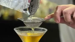mãos de um bartender coando algum liquido em uma taça (que tem alguma bebida bem amarela jã nela)