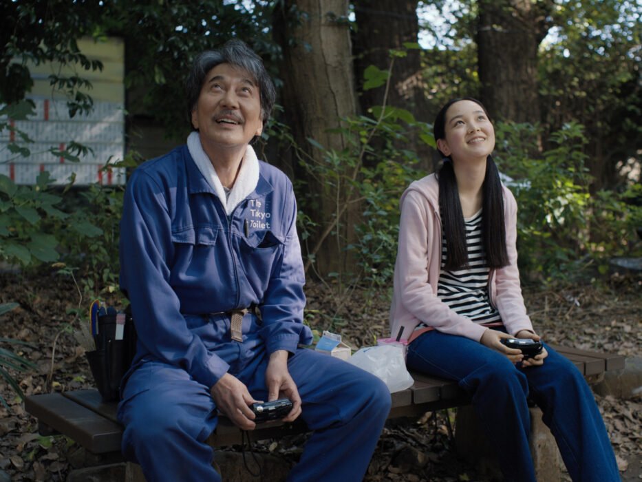 japones de meia idade, com uniforme de limpeza, sentado em um banco ao lado de uma jovem de maria-chiquinha, ambos contemplam algo acima deles, sorrindo e segurando máquinas fotográficas nas mãos. 