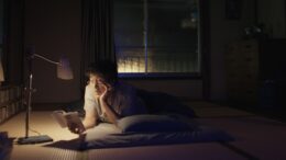 imagem de um homem deitado em tatame de um quarto tradiconal japonês, no escuro com apenas uma luminária, lendo um livro.