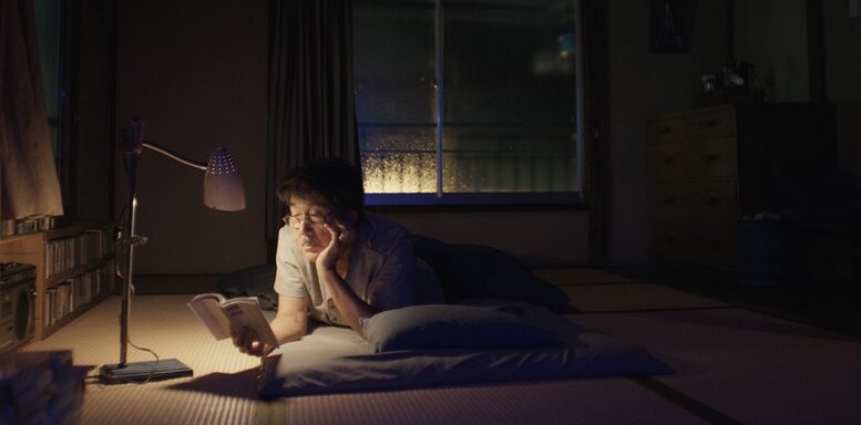 imagem de um homem deitado em tatame de um quarto tradiconal japonês, no escuro com apenas uma luminária, lendo um livro.