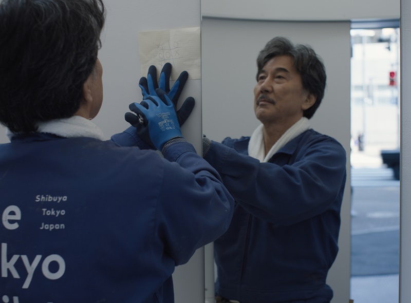 japones de meia idade, com uniforme de limpeza, segurando contra a parede um papel com o desenho de um jogo da velha. 