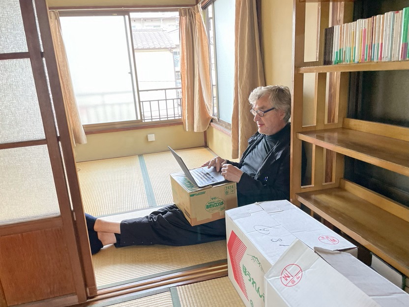 O diretor Wim Wenders sentado no chão no quarto que do protagonista do filme. ele trabalha com um notebook, apoiado sobre uma caixa de papelao sobre suas pernas esticadas. 