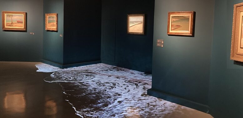 sala de exposição com paredes verde escuras, alguns quadros e a projeção de ondas do mar na area, no são da sala.