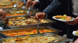 imagem de algumas mãos se servindo de comida em um buffet 'tipo quilo', com vários recipientes.