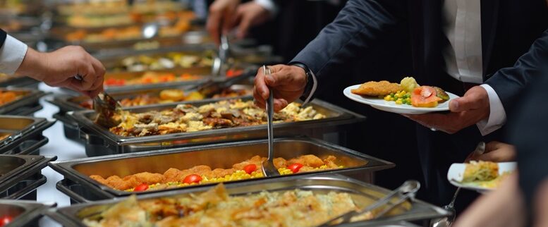 imagem de algumas mãos se servindo de comida em um buffet 'tipo quilo', com vários recipientes.