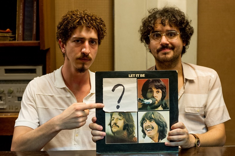 imagem dos dois personagens mostrando a capa do LP Let it Be, dos Beatles, com a foto de Lennon coberta por um papel e um ponto de interrogação dele. 