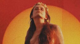 imagem de Fernanda Young em laranja e avermelhado, ela tem cabelos longos divididos e cobrindo os seios, uma tiada de flores e olhos fechados sorrindo com a cabeça voltada um pouco para cima.