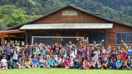 grupo grande de crianças e monitores sentados em gramado em frente a uma casa com a placa informando "Acampamento Paiol Grande"