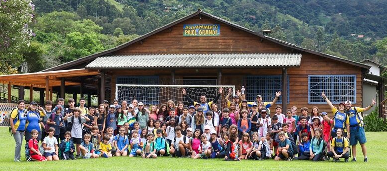 grupo grande de crianças e monitores sentados em gramado em frente a uma casa com a placa informando "Acampamento Paiol Grande"