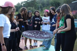 grupo de meninas em um jogo em que seguram um grande disco com bolinhas por alças, tentando deslocar as bolinhas para o centro do círculo.