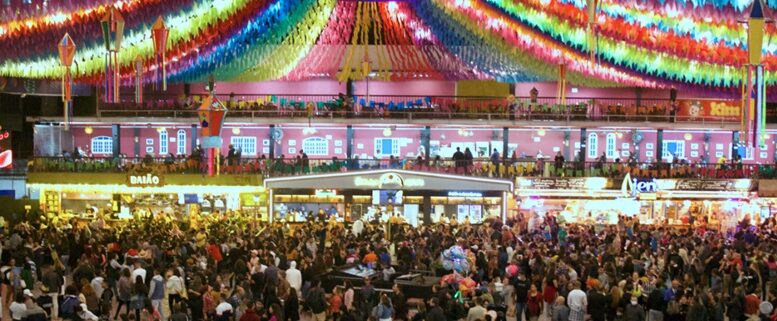 multidão na festa junina, e no teto faixas coloridas enfeitam o local