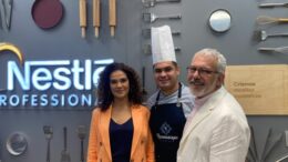 em pé, em frente a uma parede na Nestlé, com a logo da empresa e alguns utensílios de cozinha como decoração, posam Helen, Felipe e João.