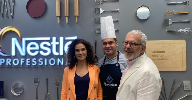 em pé, em frente a uma parede na Nestlé, com a logo da empresa e alguns utensílios de cozinha como decoração, posam Helen, Felipe e João.