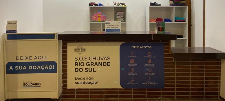 imagem de um balcão sinalizado como ponto de coleta "SOS Chuvas Rio Grande do Sul" e uma grande caixa de papelão com os mesmos informes.