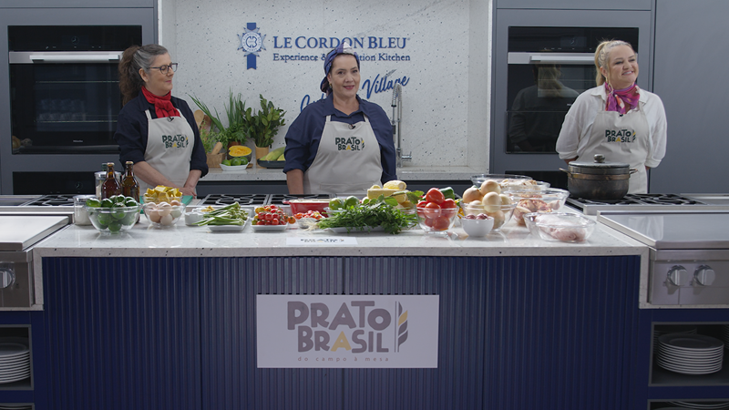 três mulheres chefs com aventais de copzinha atrás de uma bancada com ingredientes, na bancada está a logo programa Prato Brasil e na parede atrás, a logo do Le Cordon Bleu