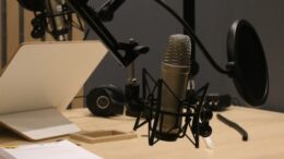 imagem de uma mesa de gravação com microfone, abafador, um notebook visto por tras e alguns papéis sobre a mesa de f´rmica clara.