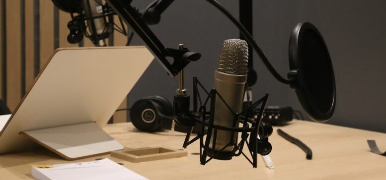 imagem de uma mesa de gravação com microfone, abafador, um notebook visto por tras e alguns papéis sobre a mesa de f´rmica clara.
