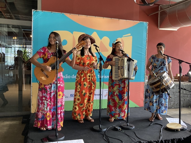 grupo musical com 4 mulheres de vestido de chita, tocando instrumentos como violão, triangulo, acordeon e um tambor especial