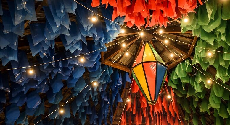 imagem de um teto de festa junina, com muitas bandeiras e um balão iluminado pendurado, tudo em tonss de vermleho verde e azul.