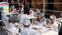 grupo de crianças com deficiencia visual sentadas à mesa com avental e touca de cozinha, com pratinhos de plástico em frente a eles.