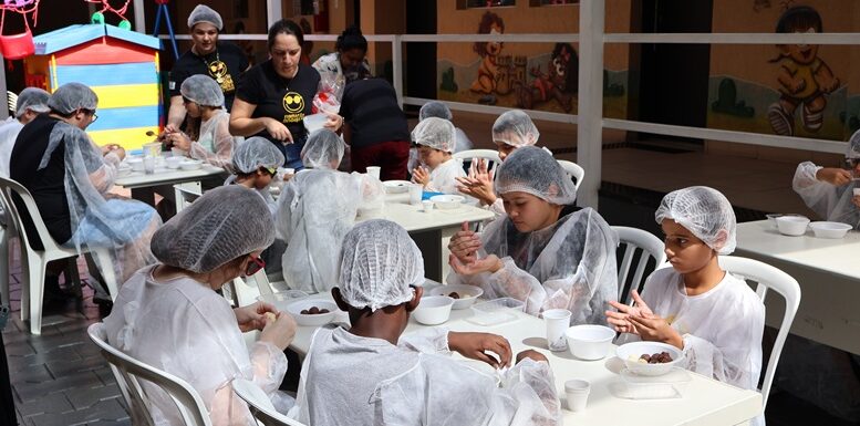 grupo de crianças com deficiencia visual sentadas à mesa com avental e touca de cozinha, com pratinhos de plástico em frente a eles.