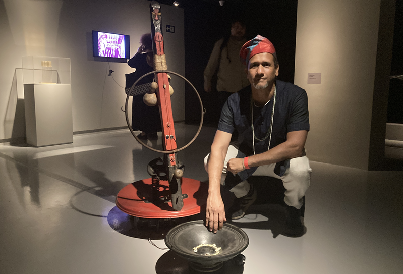 Artista agaixado ao lado da obra Exu, um totem vermelho, com elementos de metal e a frente um alto falante sem a caixa, virado para cima, com alguns búzios. 